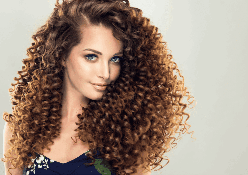 Mulher de frente com cabelo longo cacheado e brilhante em movimento, mostrando a saúde dos fios após cuidados capilares.