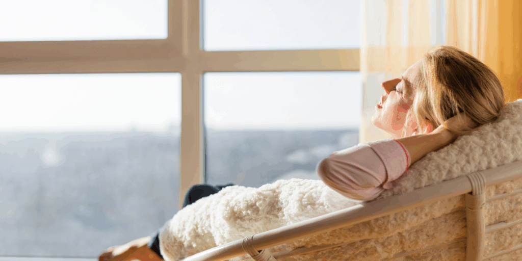  Mulher de olhos fechados, respirando fundo perto de uma janela com luz suave, praticando uma das maneiras eficientes de relaxamento.