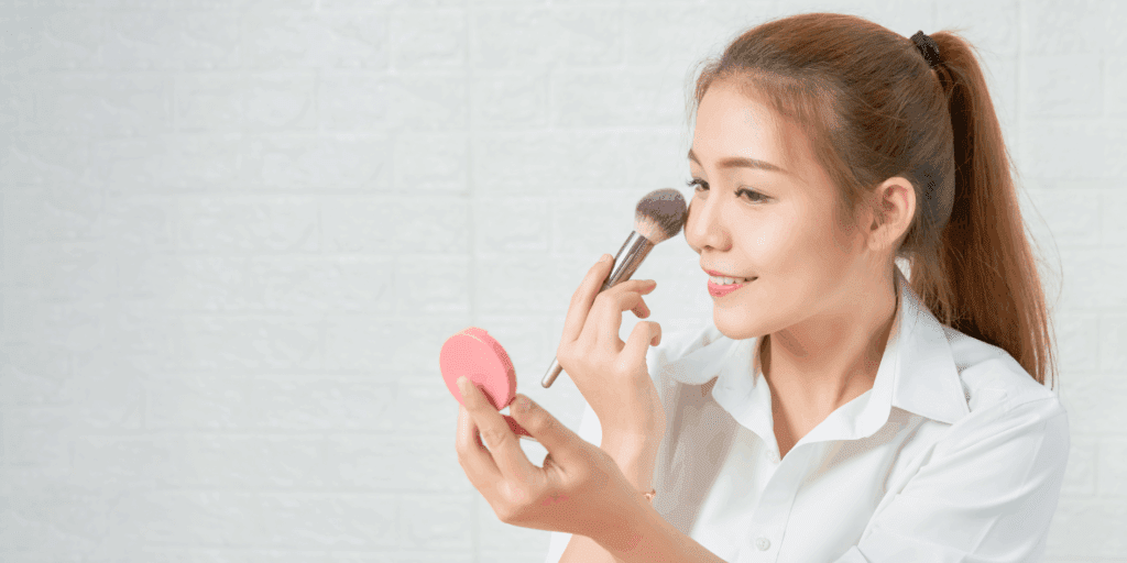 Close-up de uma mulher aplicando um produto multifuncional nas bochechas com os dedos, com um sorriso leve.