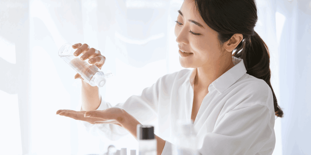 Mulher sorrindo enquanto faz sua rotina de skincare, representando os pilares de limpeza, hidratação e proteção.