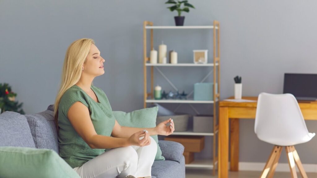 Uma mulher sentada em posição de lótus (meditação), mas com uma expressão serena e roupas confortáveis, em um ambiente de casa normal (não no topo de uma montanha inalcançável). Passa a ideia de "meditação possível".