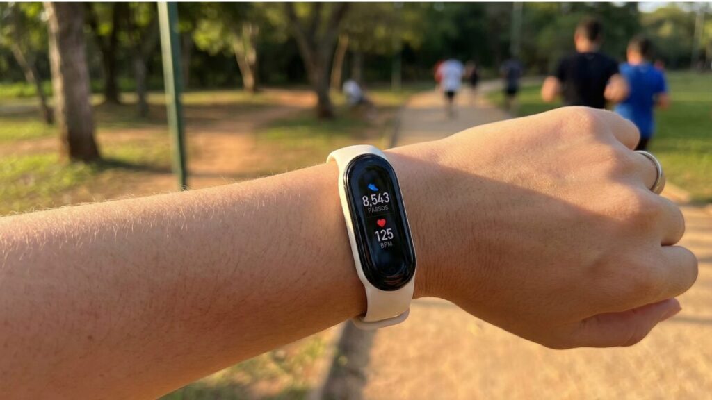 Um pulso feminino usando uma Mi Band (pulseira preta fina) mostrando a tela com o contador de passos ou batimentos cardíacos. Fundo de treino ou caminhada.
