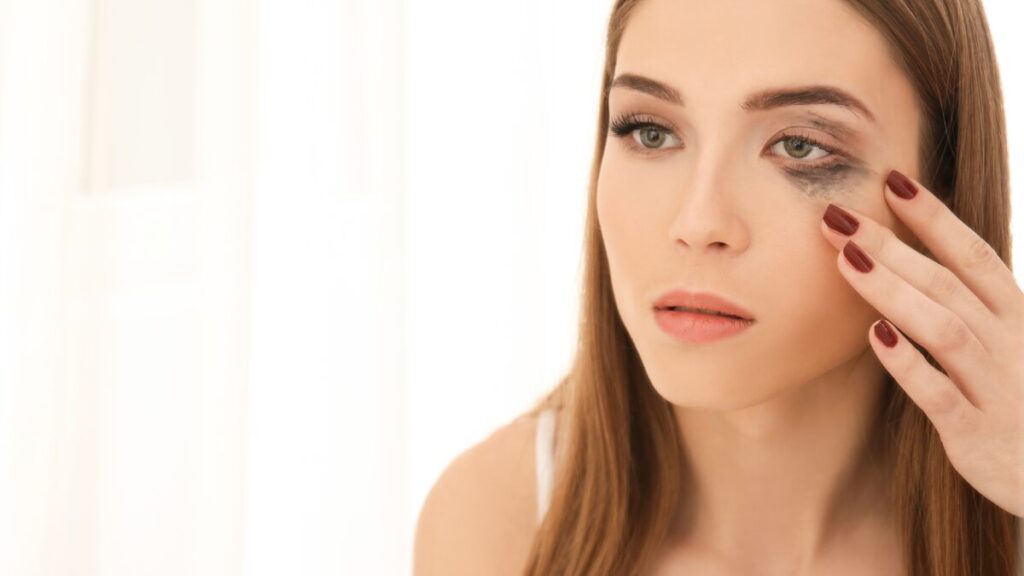 Um close-up dos olhos de uma mulher olhando para cima, mostrando uma mancha preta (borrão de rímel) logo abaixo dos cílios inferiores. A expressão deve ser de chateação, ilustrando o famoso "olho de panda"