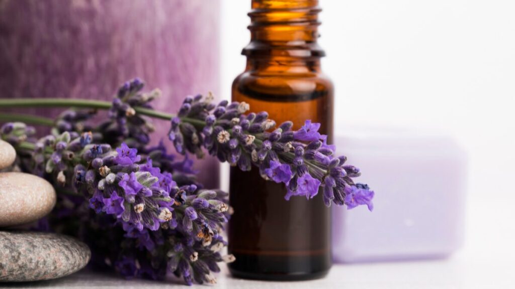 Uma foto relaxante de um frasquinho de óleo essencial de lavanda aberto, com algumas flores de lavanda roxas ao lado, em cima de uma roupa de cama branca ou uma mesa de madeira clara. A luz deve ser suave.