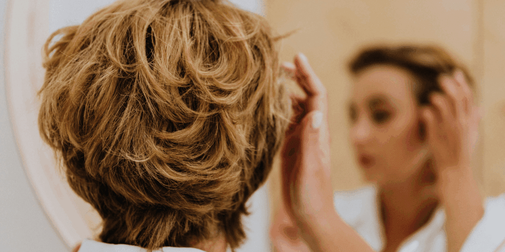 Uma foto de uma mulher loira (com luzes) no chuveiro, com a mão na cabeça, com uma expressão levemente "preocupada" ou "pensativa" sobre o cabelo. A imagem deve capturar a "dúvida" e o "medo" de usar um produto que pode ressecar os fios claros.